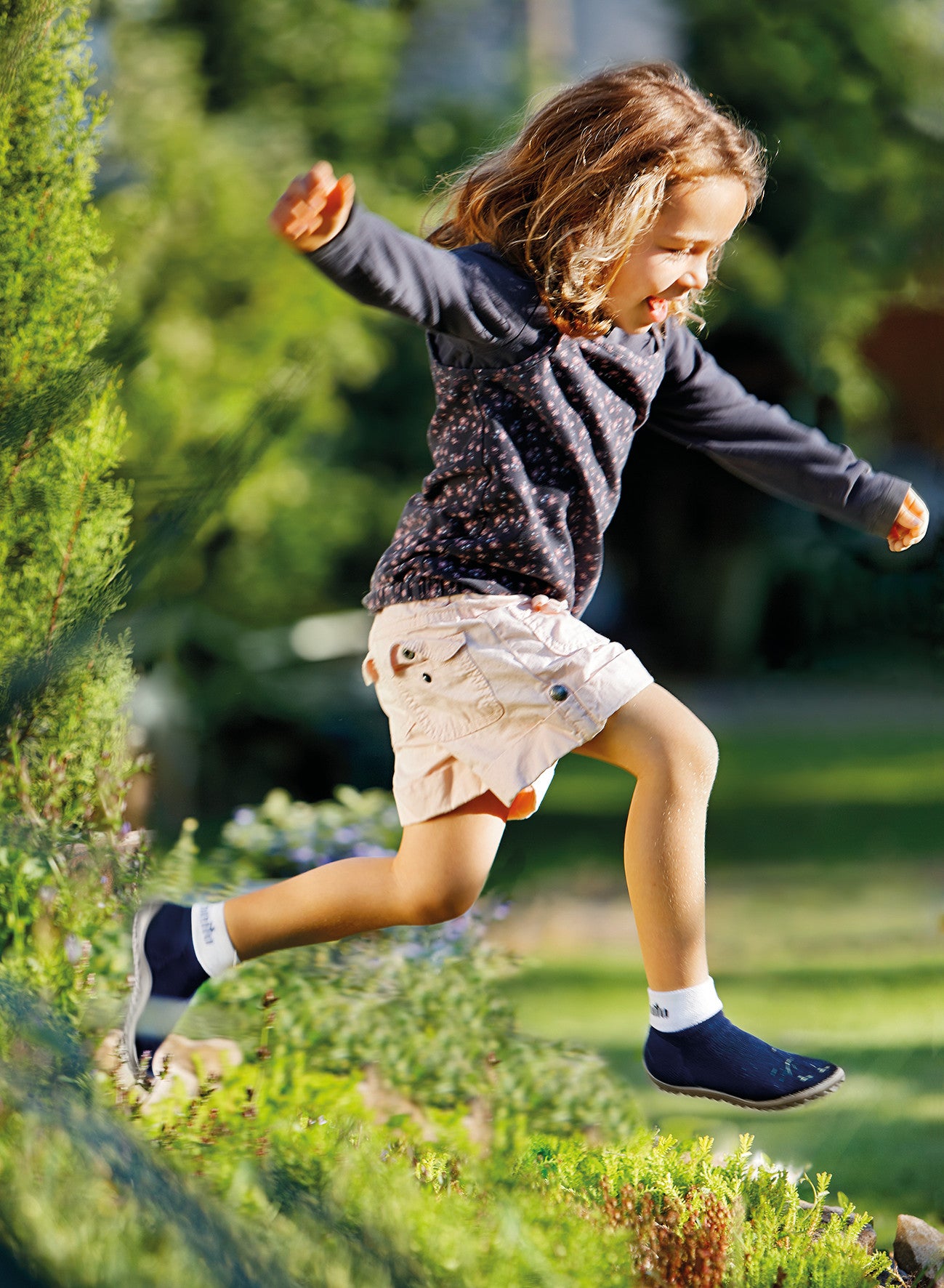 Leguanito Barfußschuhe für Kinder von Leguano in Blau – Sockenschuhe, getragen von einem Kind, das sich mitten im Sprung über eine grüne Hecke befindet, erhältlich im barfusswelteb.com Barfußschuhe Onlineshop