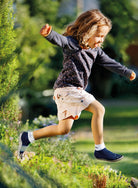 Leguanito Barfußschuhe für Kinder von Leguano in Blau – Sockenschuhe, getragen von einem Kind, das sich mitten im Sprung über eine grüne Hecke befindet, erhältlich im barfusswelteb.com Barfußschuhe Onlineshop